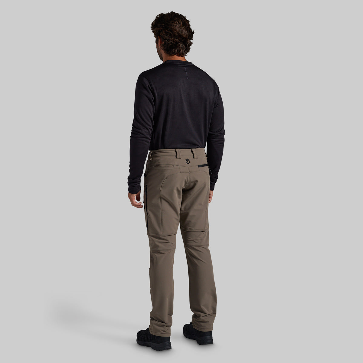 A man viewed from the back wearing a black long-sleeve shirt and grey technical pants, standing against a solid light gray studio background.