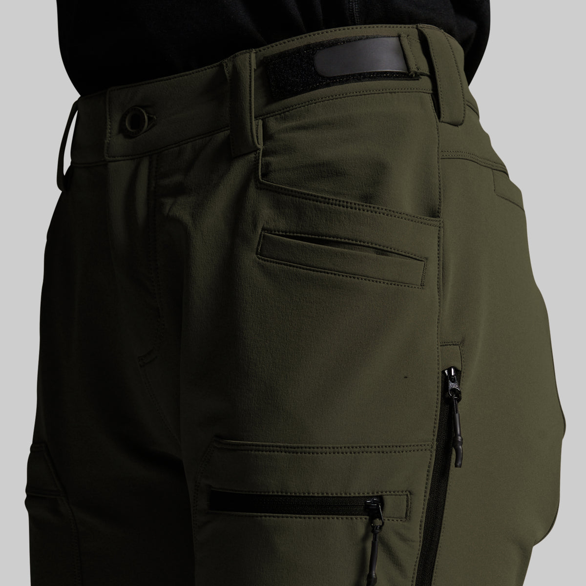 Close-up of olive green outdoor pants showing the side pocket, belt loop, and zippered cargo pocket on a neutral gray background.