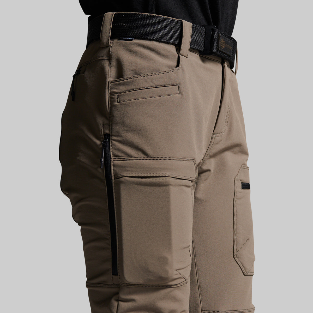 Close-up of a person wearing grey outdoor cargo pants with black zippers, belt loops, and a black belt, shown against a light gray studio background.