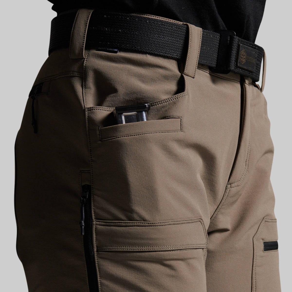 Close-up of a person wearing grey outdoor cargo pants with a black belt, showing side pockets and a zippered compartment.