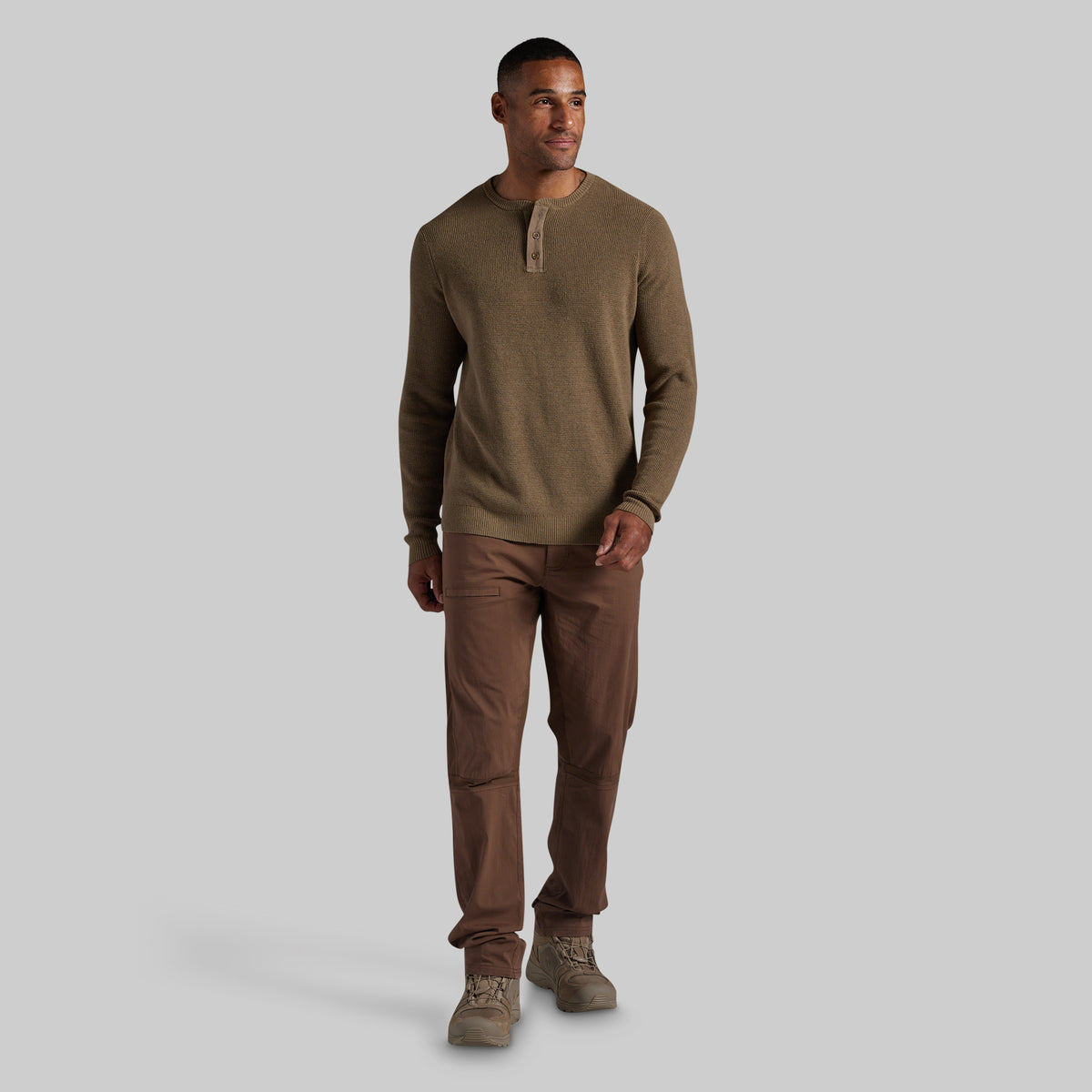 A man modeling a long-sleeve henley shirt in olive brown and brown pants against a solid gray studio background.