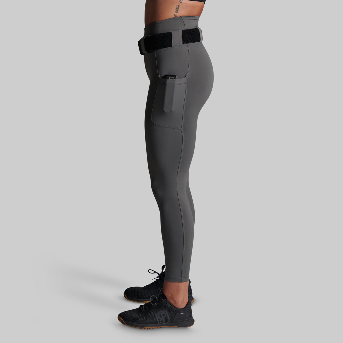 Side view of a model wearing dark gray high-waisted athletic leggings with a black belt and side pocket, paired with black athletic shoes, against a solid gray background.