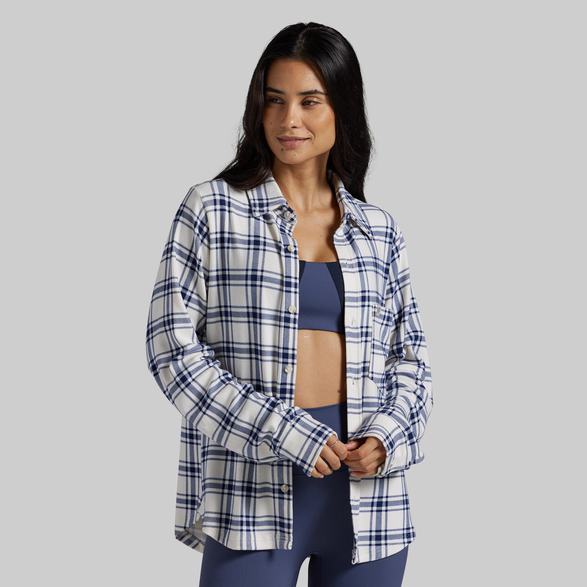 A woman modeling a long-sleeve button-up plaid shirt in navy, white, and light blue over a navy sports bra and matching leggings, set against a solid gray studio background.