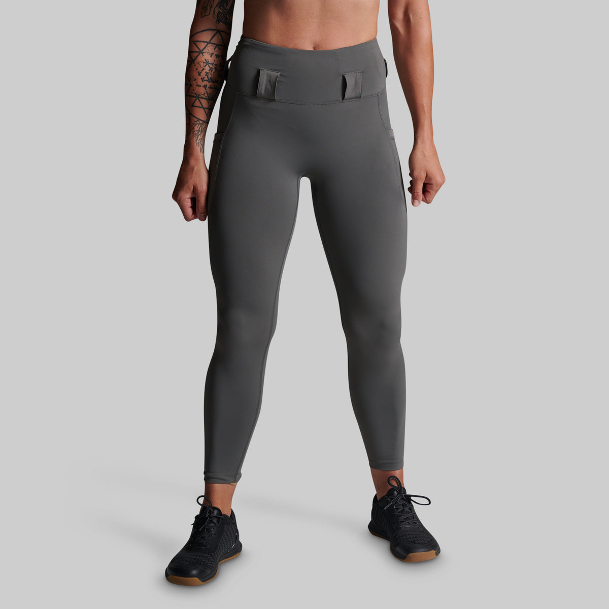 A model from the waist down wearing dark gray high-waisted athletic leggings with belt loops and black athletic shoes, set against a solid light gray background.