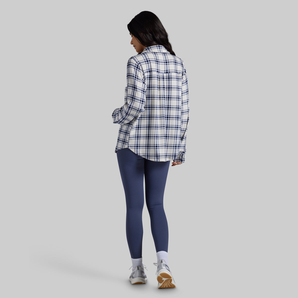 A woman is shown from behind wearing a white long-sleeve plaid button-up shirt with thin navy stripes, paired with navy blue leggings, white socks, and gray sneakers, against a solid gray background.