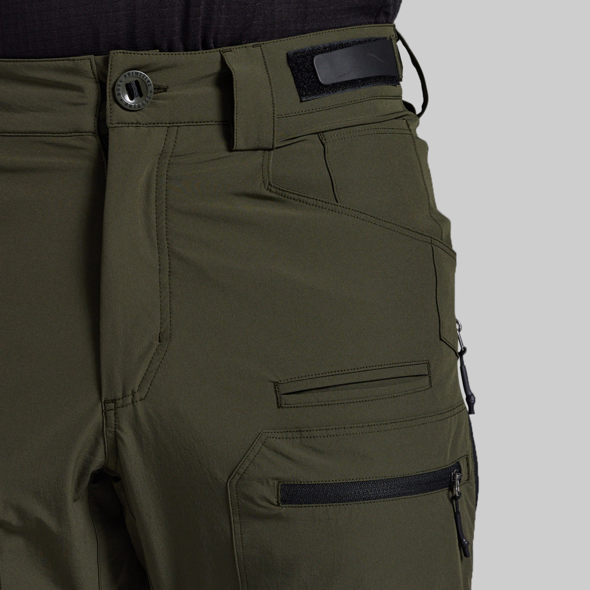 Close-up of olive green outdoor pants showing the waistband with a button closure, belt loop, and side cargo pocket with a zippered pocket on a light gray background.