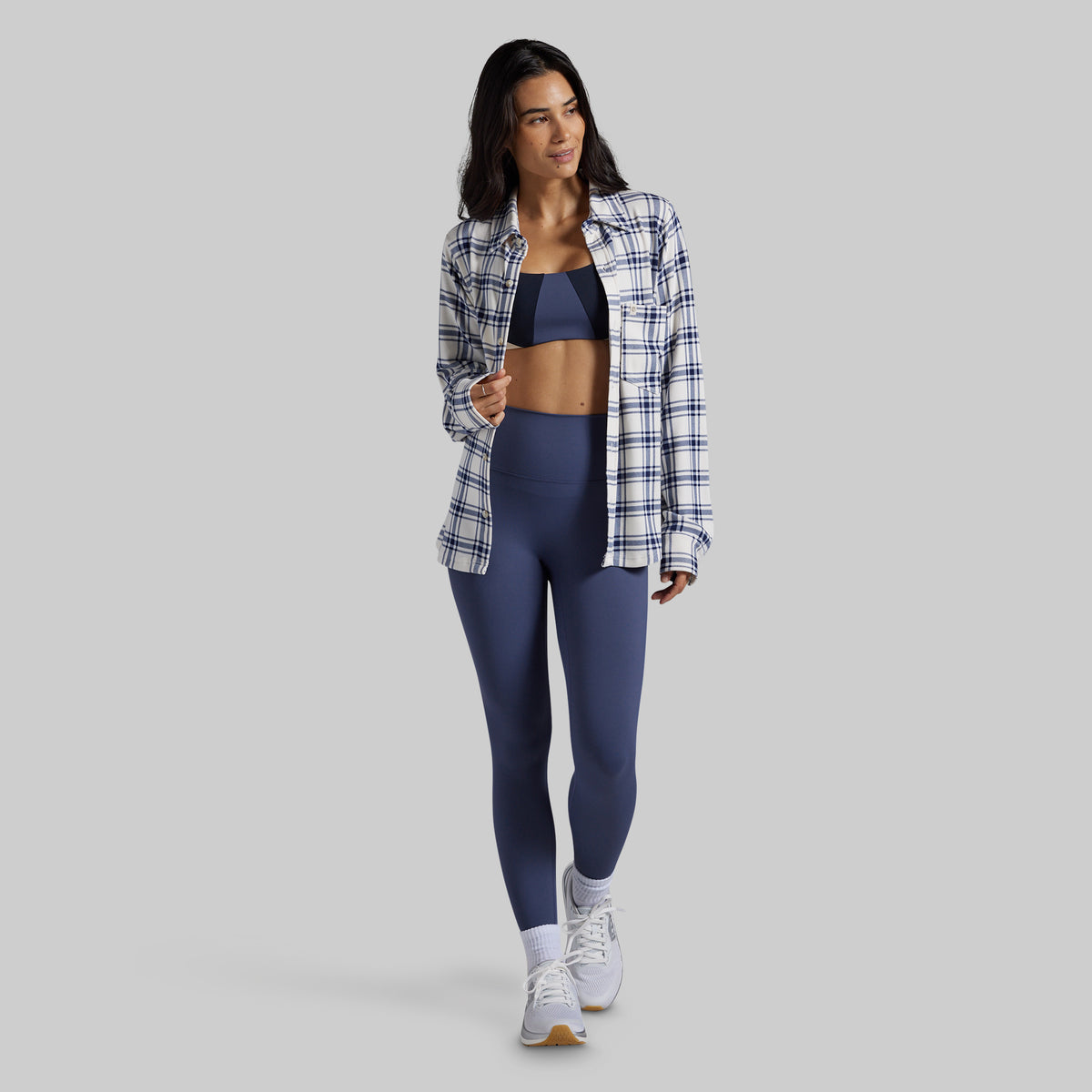A woman modeling a matching athletic set consisting of a navy blue sports bra and high-waisted leggings, styled with an open navy and white plaid flannel shirt, white socks, and white sneakers against a solid gray background.