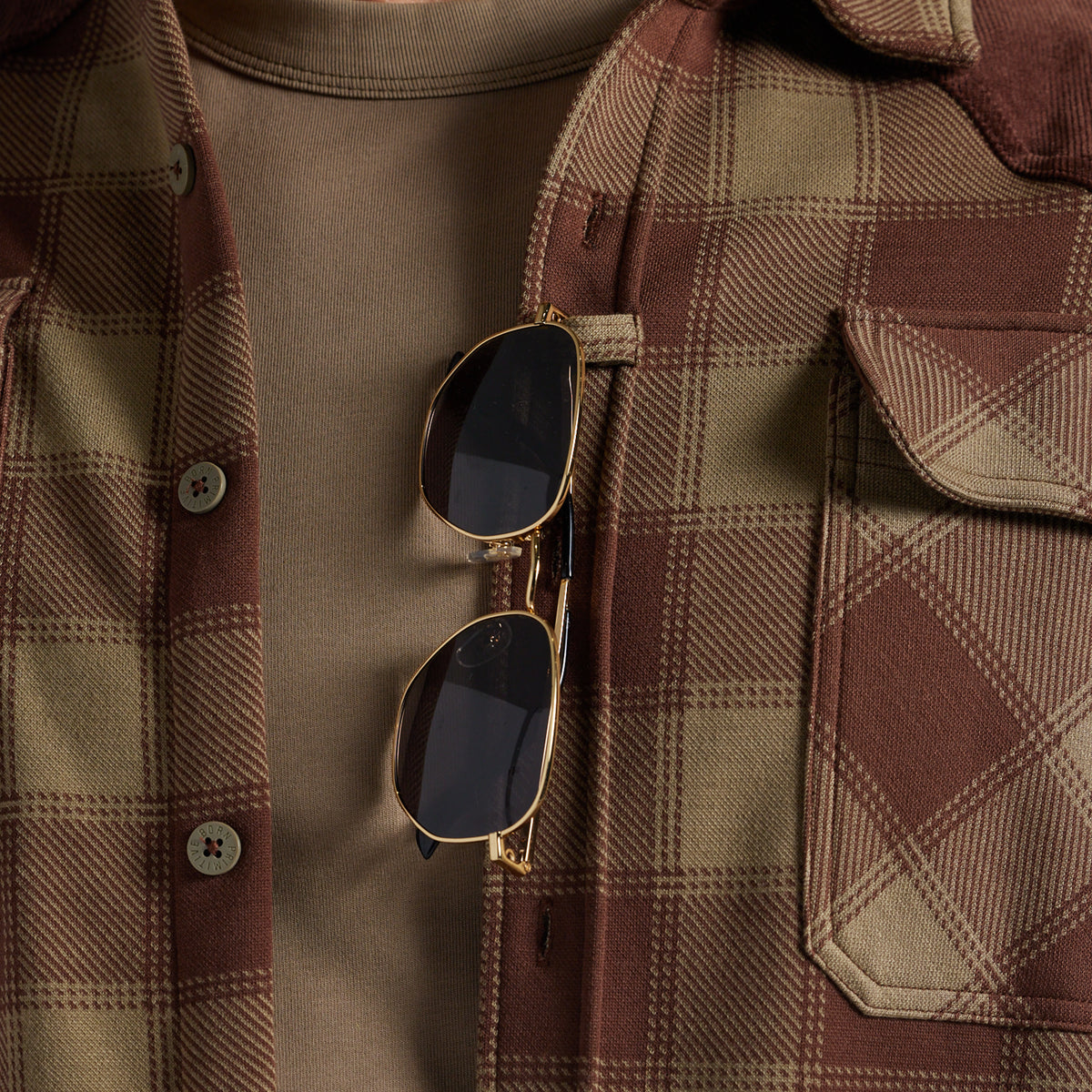 Close-up of a person wearing a brown and tan plaid flannel shirt over a taupe t-shirt, with gold-framed sunglasses hanging from the placket.