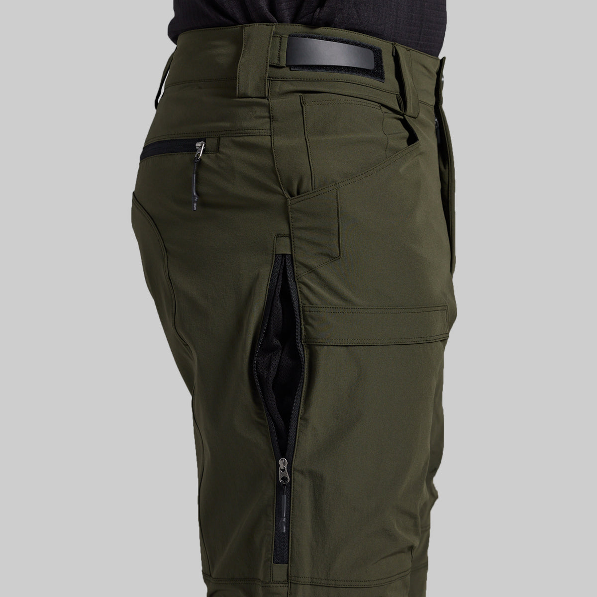 Close-up of olive green cargo pants with multiple zippered pockets, shown on a model from the waist down against a light gray background.