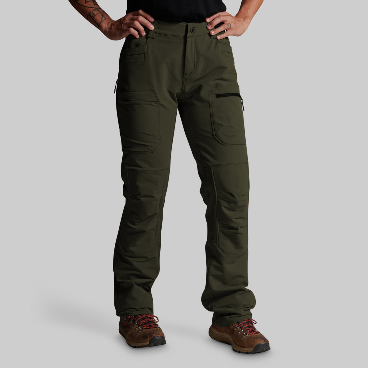 A person wearing dark olive green cargo pants with brown hiking boots, shown from the waist down against a light gray studio background.