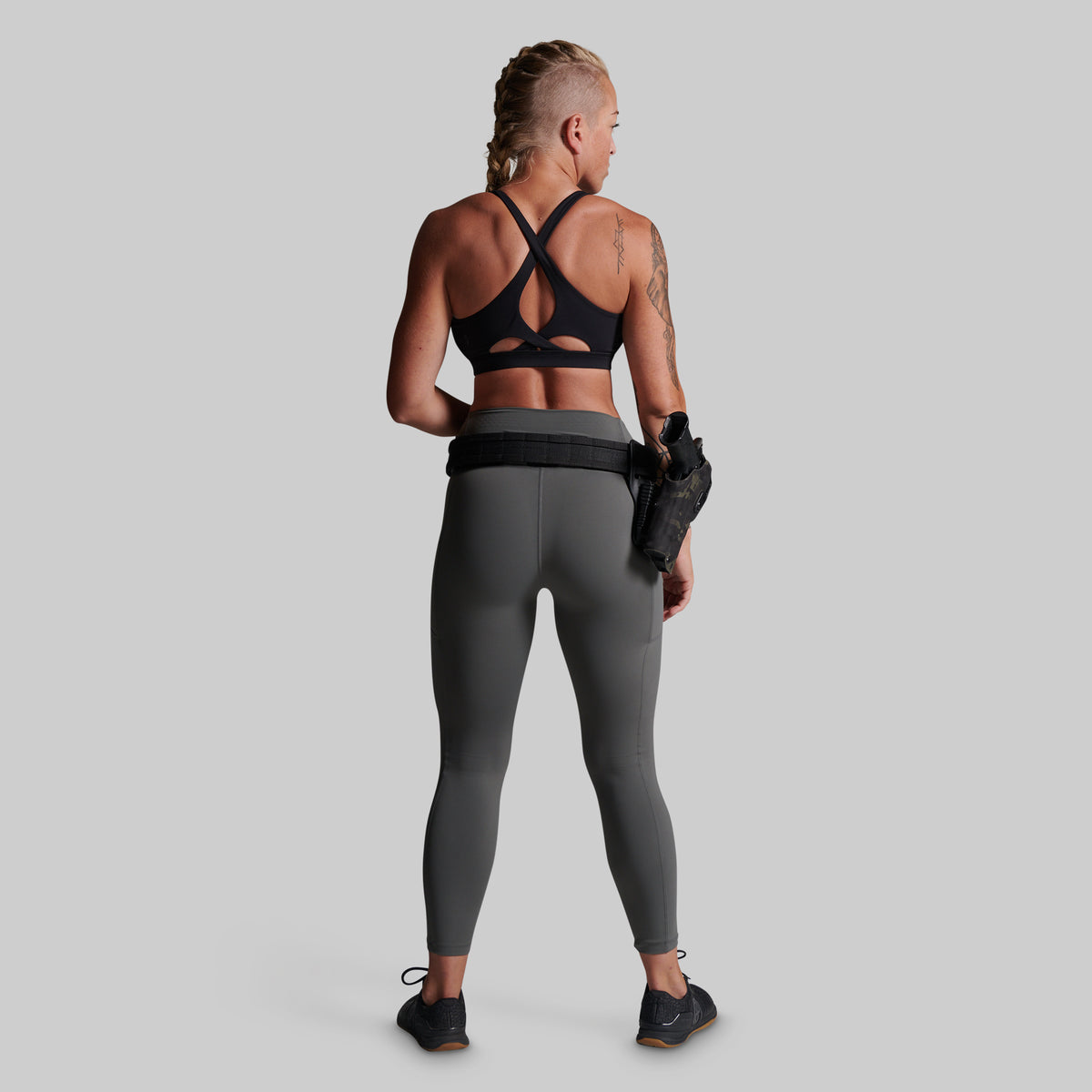 A woman shown from the back wearing dark gray full-length leggings and a black sports bra, standing against a light gray studio background. She is wearing a black belt with a holster