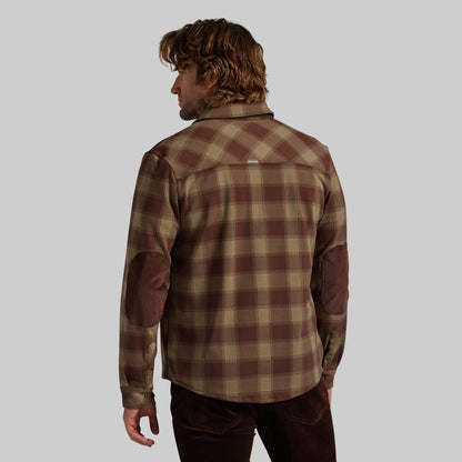 A man shown from the back wearing a brown and beige plaid flannel shirt with brown corduroy elbow patches, set against a solid gray background.