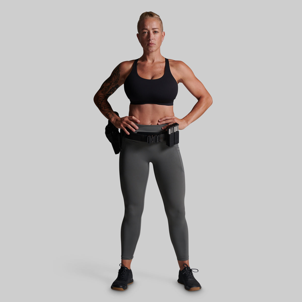 A woman wearing a black sports bra, dark gray full-length leggings with a utility belt and holster, and black athletic shoes.
