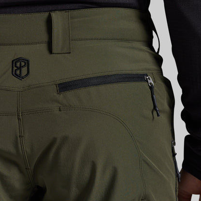 Close-up of olive green outdoor pants showing a zippered pocket, belt loop, and a black embroidered logo on the side, worn with a black long-sleeve top against a light gray background.
