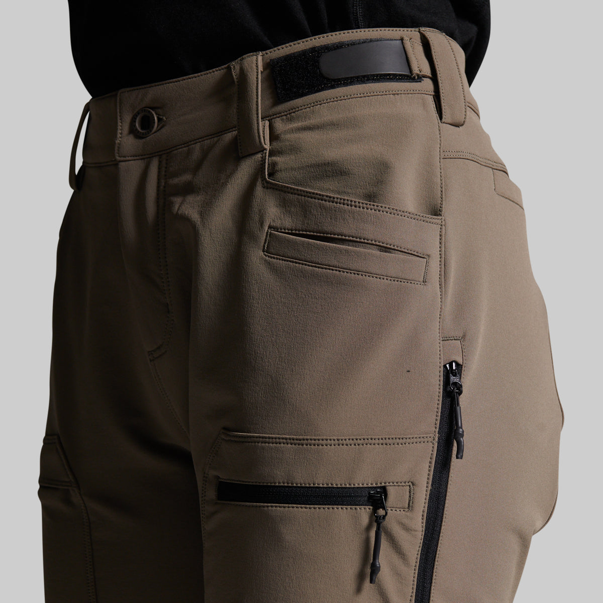 Close-up of a person wearing grey cargo pants with black zippers, belt loops, and a black waistband, shown against a light gray studio background.