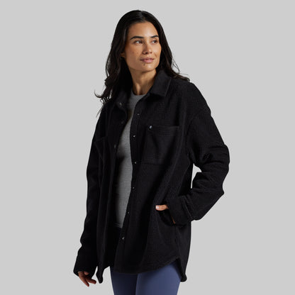 A woman modeling a black textured fleece button-up overshirt over a gray t-shirt and blue leggings, photographed against a solid gray studio background.