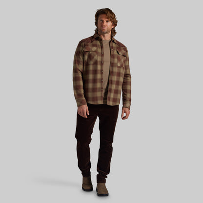 A man modeling a brown and beige plaid flannel shirt over a tan undershirt, paired with dark brown pants and brown boots, against a solid gray background.