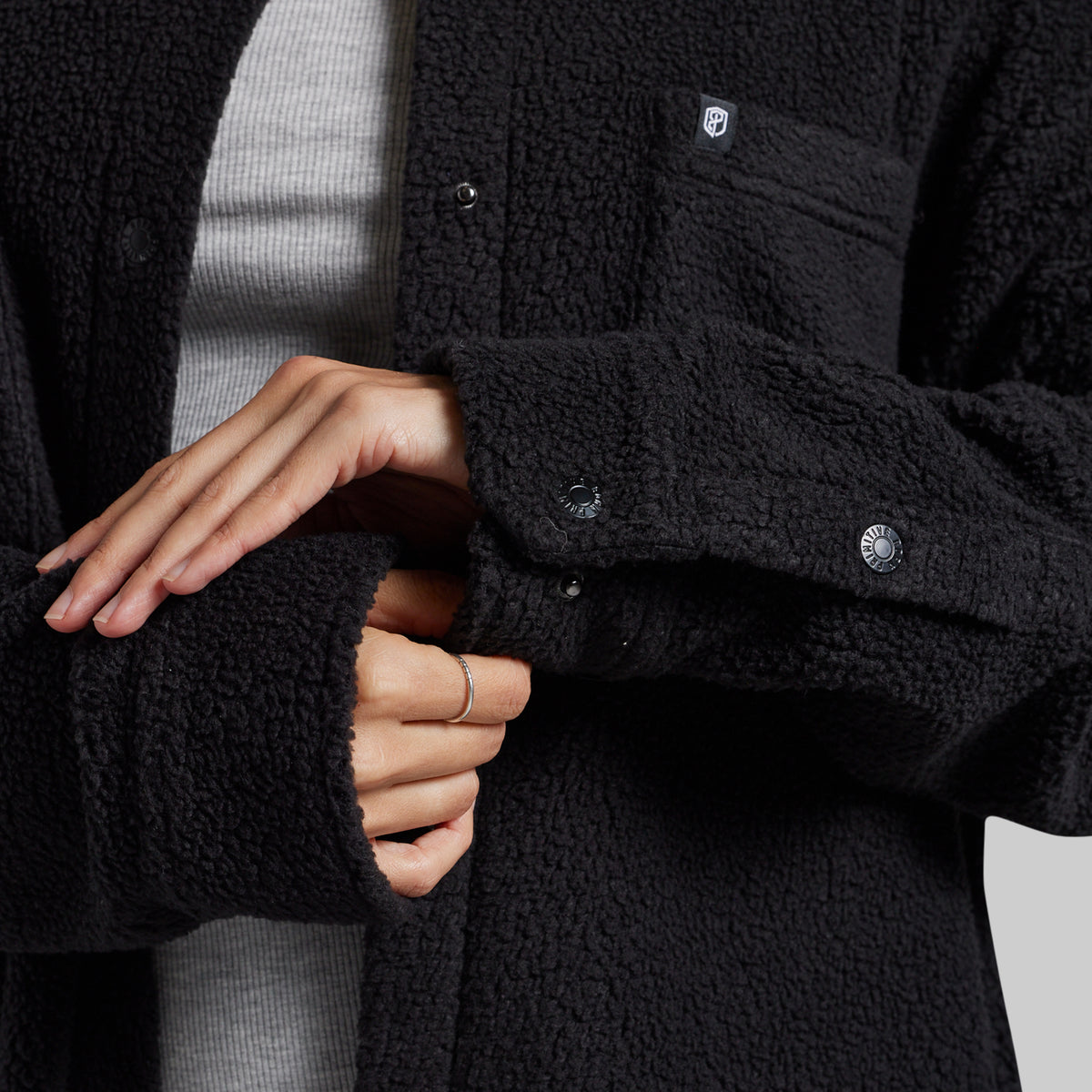 Close-up of a person wearing a black sherpa fleece button-front jacket over a light gray ribbed top, with both hands visible adjusting the sleeve cuff.