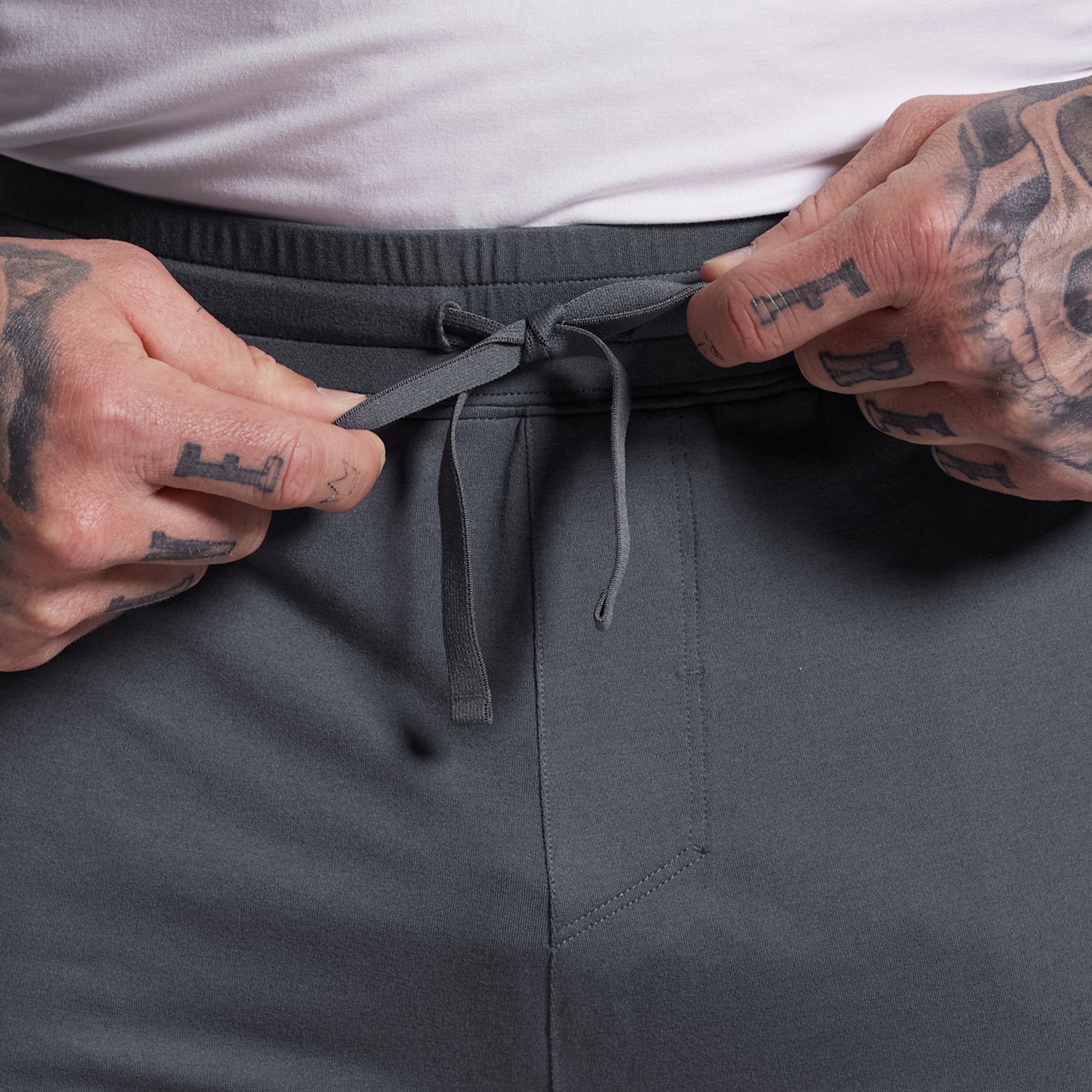 Person adjusting the drawstring of gray shorts with a tattooed hand.