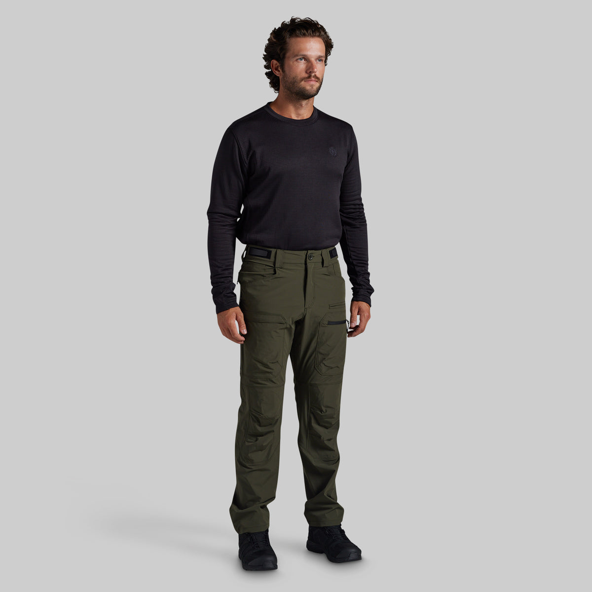 A man modeling a black long-sleeve crewneck shirt and olive green outdoor pants against a solid gray background.