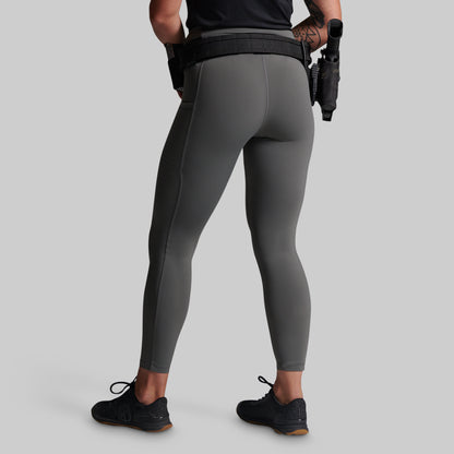 Rear view of a model wearing dark gray full-length athletic leggings with a black waist belt and black sneakers, standing against a solid light gray background.