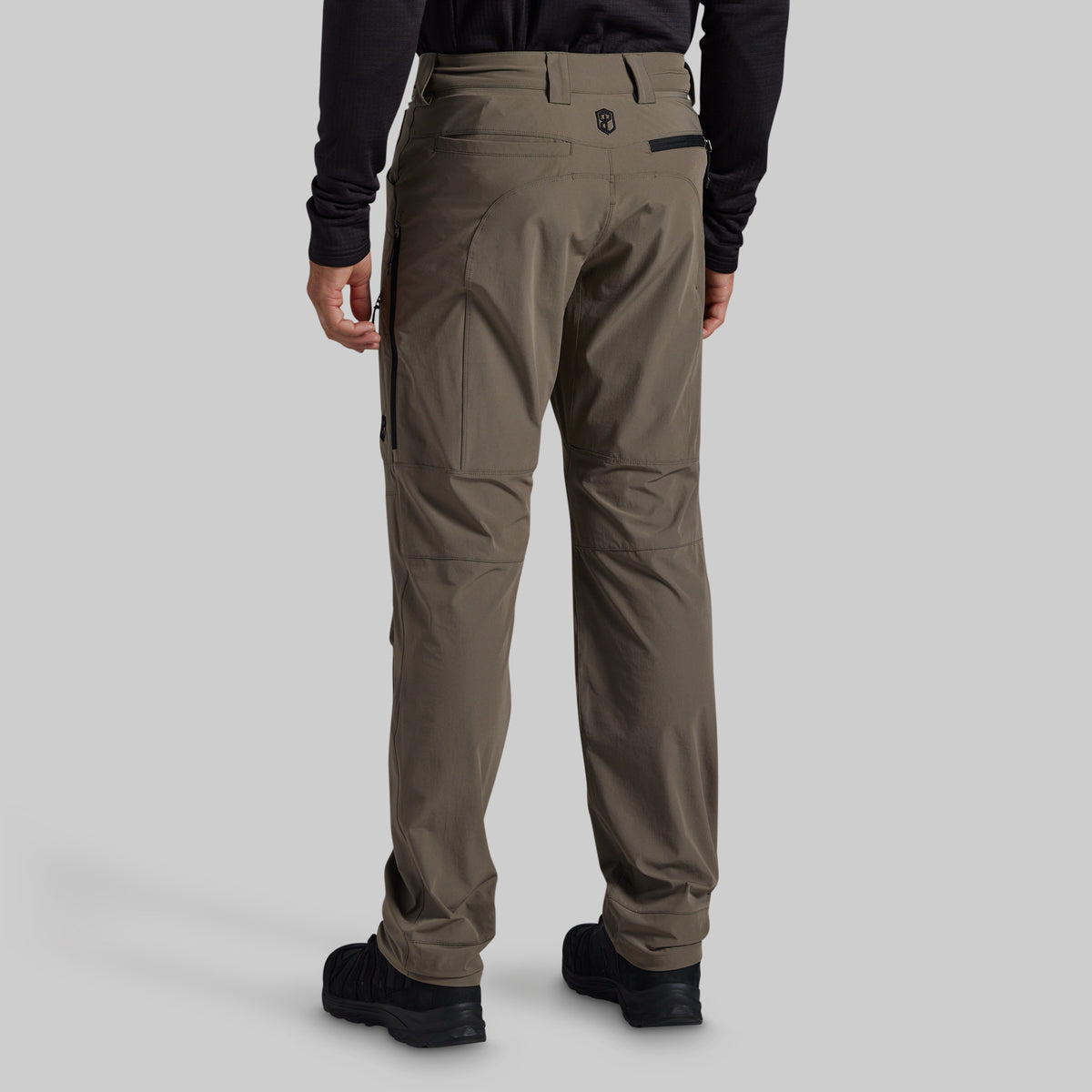 Rear view of a person wearing grey technical cargo pants with black shoes and a black long-sleeve top, shown against a light gray studio background.