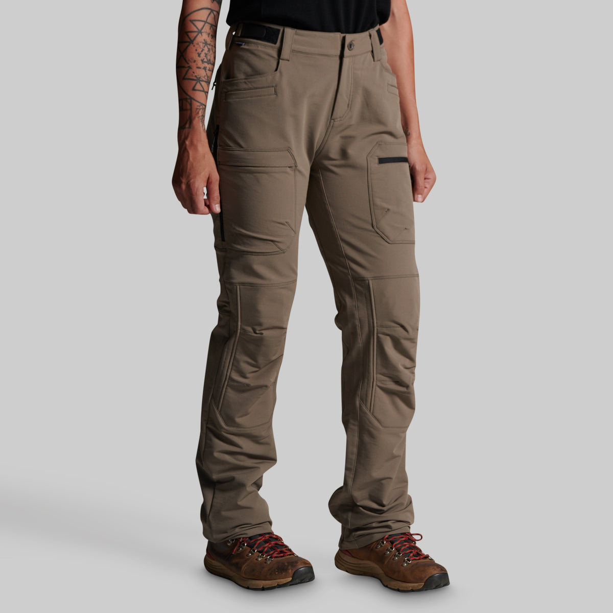 A person wearing grey cargo outdoor pants with a black belt, paired with brown hiking boots, shown from the waist down against a light gray studio background.