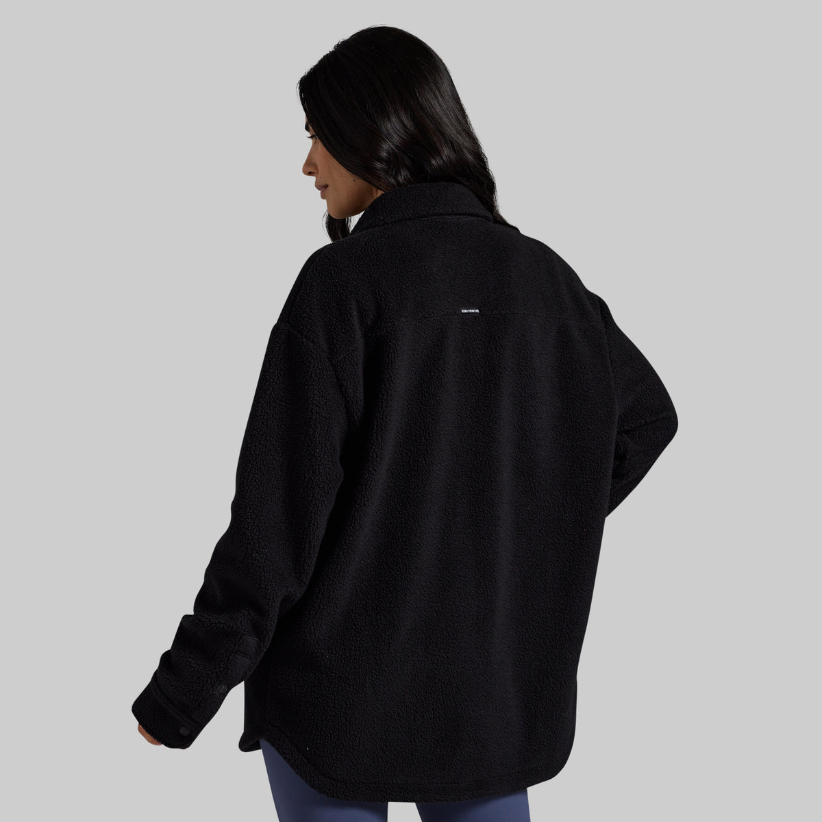 A woman shown from the back wearing an oversized black fleece jacket against a solid light gray background.
