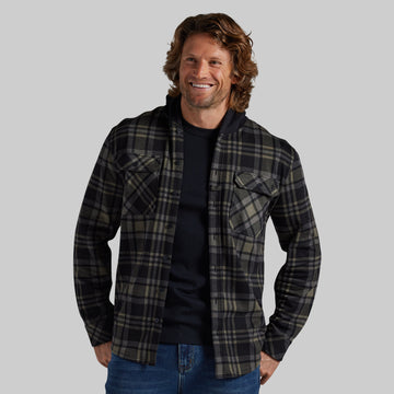 A man wearing a black and green plaid button-up flannel shirt over a black t-shirt, photographed against a solid gray studio background.