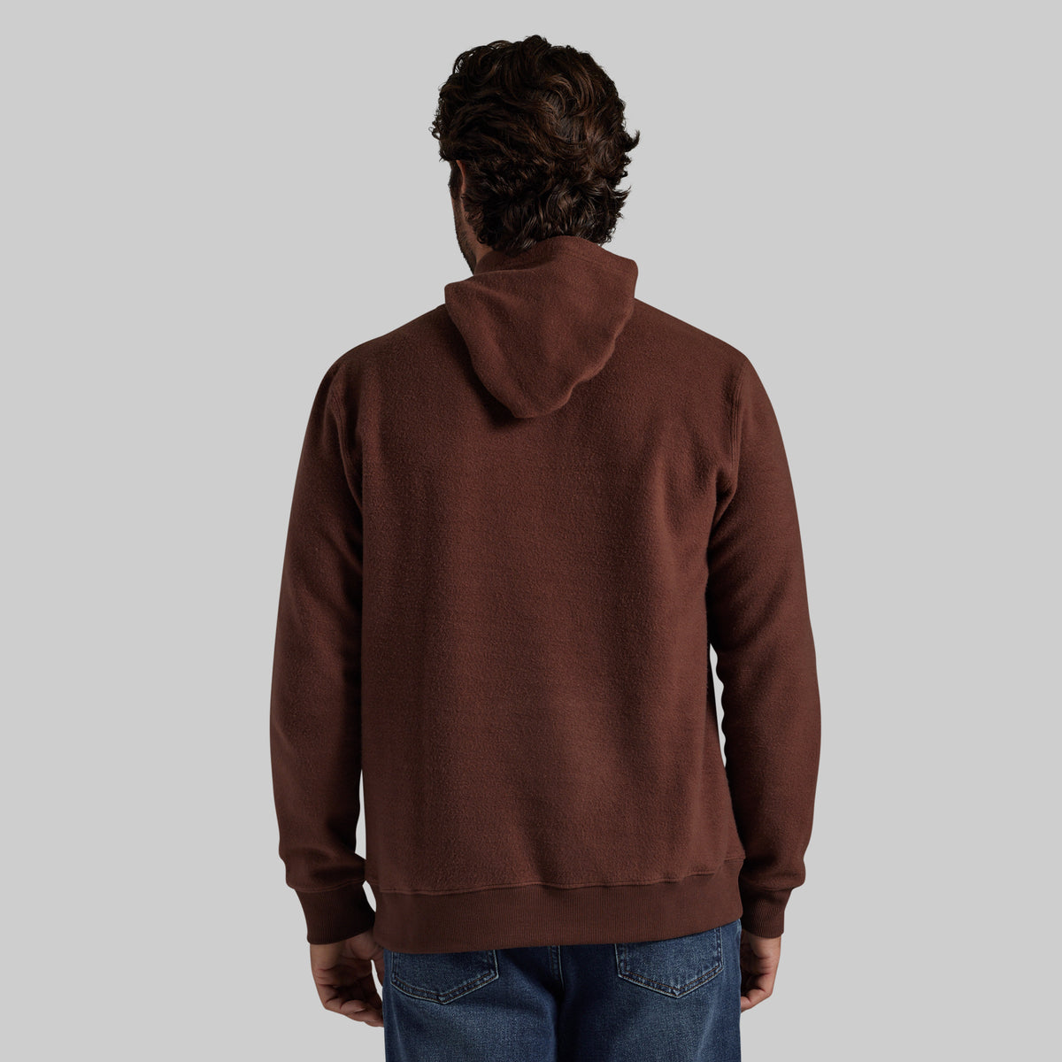 A man viewed from behind wearing a dark brown pullover hoodie and blue jeans, set against a solid light gray background.