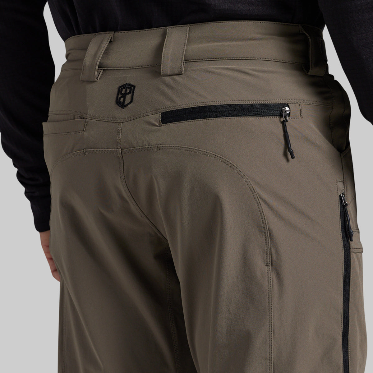 Close-up back view of a person wearing grey technical pants with a black zippered pocket and a small shield logo on the upper back, paired with a black long-sleeve top against a light gray studio background.