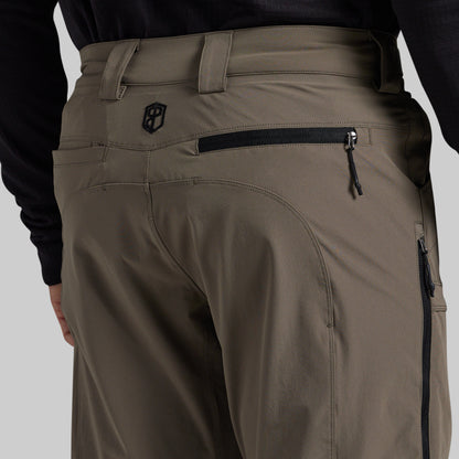 Close-up back view of a person wearing grey technical pants with a black zippered pocket and a small shield logo on the upper back, paired with a black long-sleeve top against a light gray studio background.