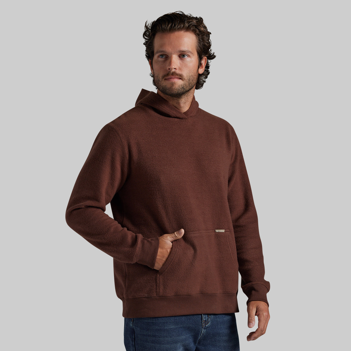 A man wearing a brown pullover hoodie with a front kangaroo pocket, paired with blue jeans, standing against a solid gray background.