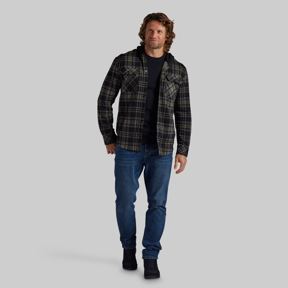 A man modeling a dark green and black plaid flannel shirt over a black undershirt, paired with blue jeans and black boots, against a solid gray background.