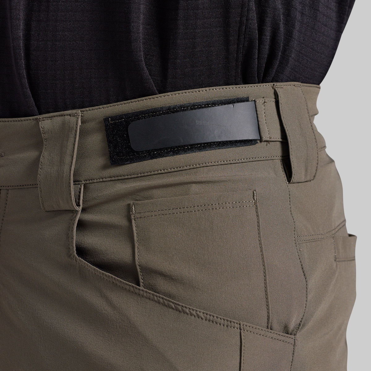 Close-up of the back waistband and pocket area of grey cargo pants, showing a black hook-and-loop patch on the belt loop, worn with a dark textured top against a light gray background.