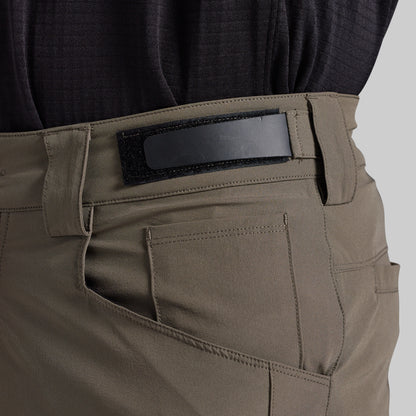 Close-up of the back waistband and pocket area of grey cargo pants, showing a black hook-and-loop patch on the belt loop, worn with a dark textured top against a light gray background.