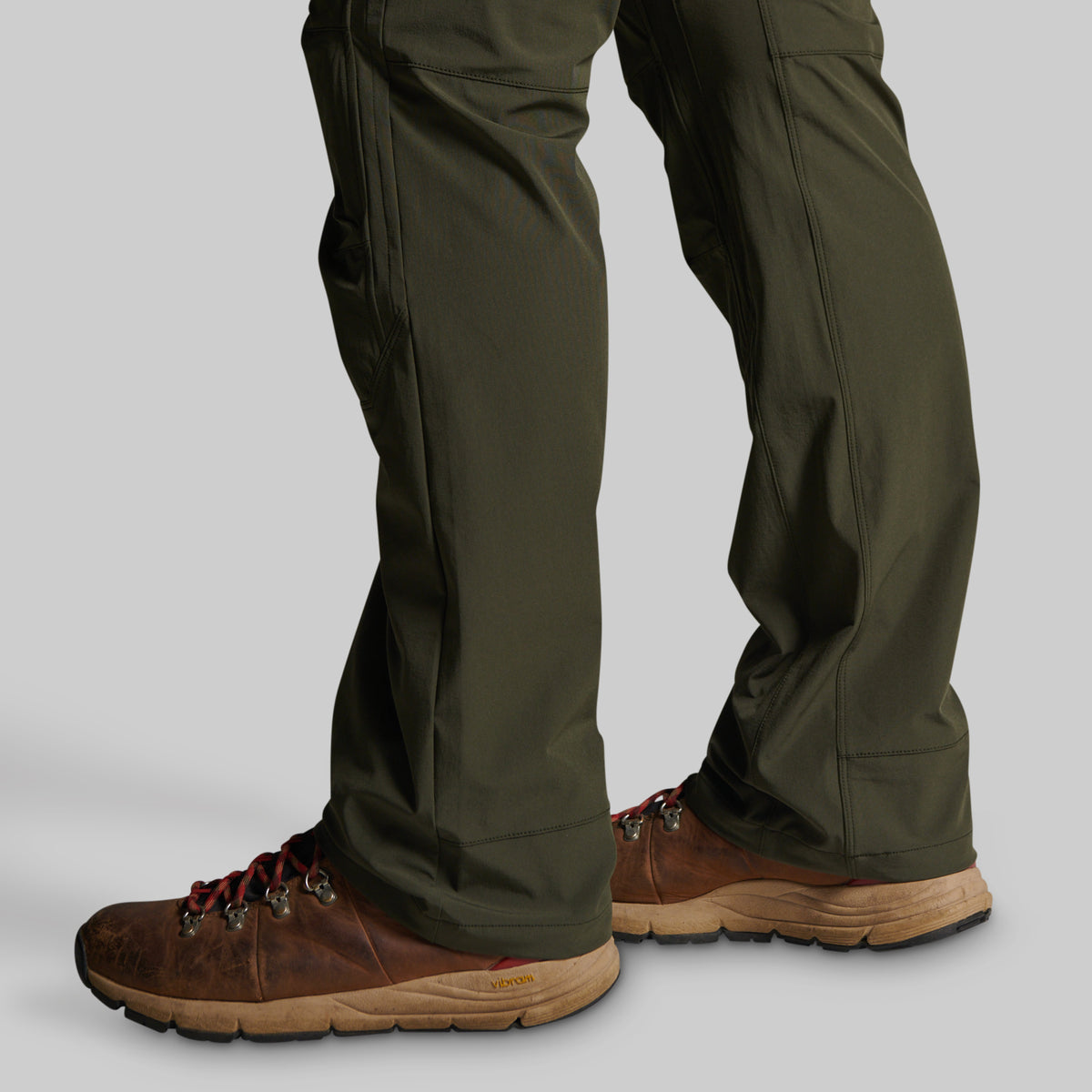 Close-up of a person's legs wearing olive green pants and brown hiking boots against a plain light gray studio background.