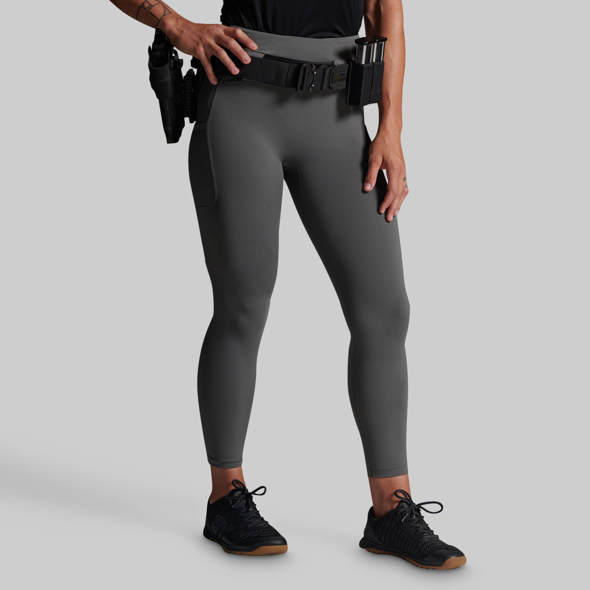 A model wearing dark gray full-length athletic leggings with a black utility belt and black tactical shoes, shown from the waist down against a solid gray background.
