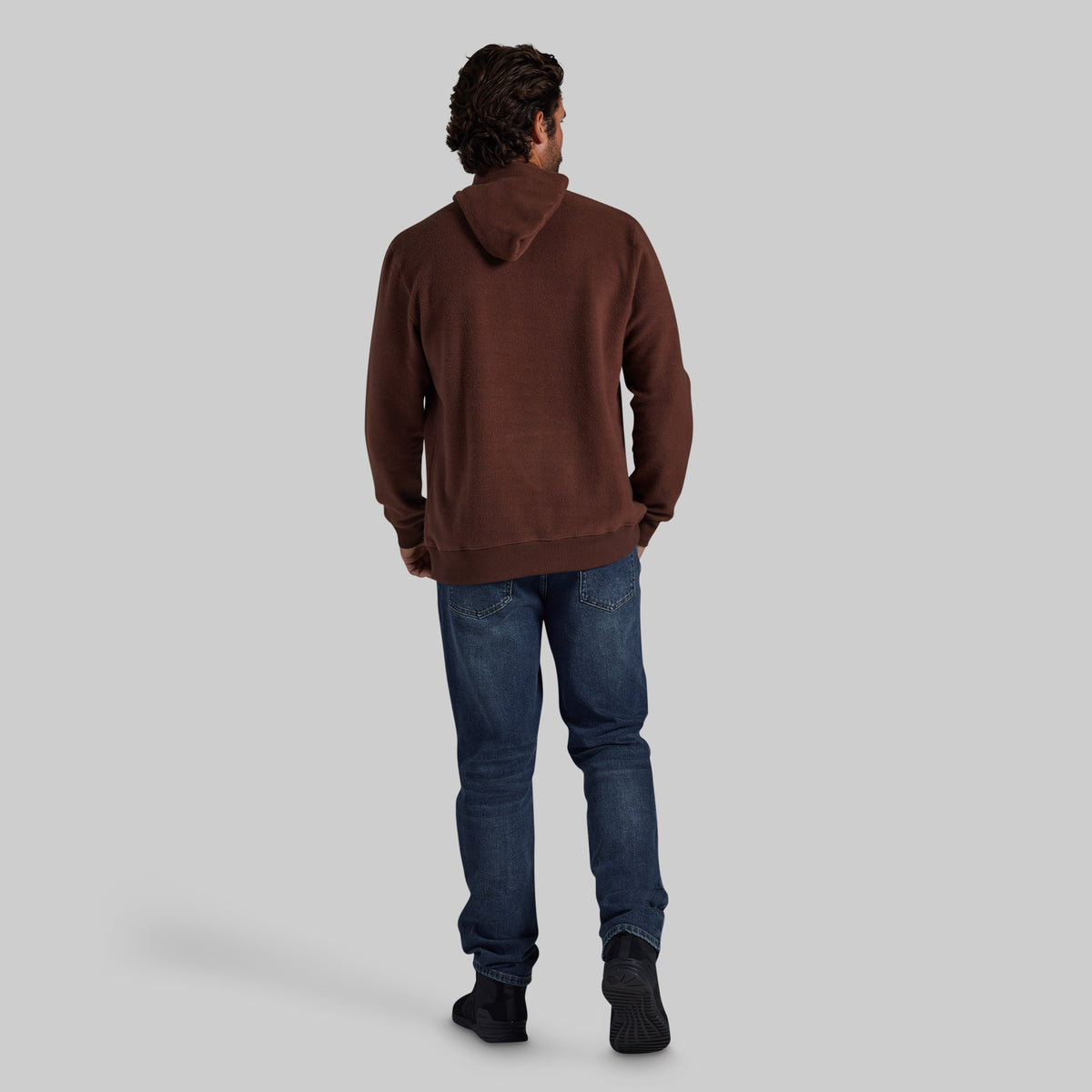 A man viewed from behind wearing a brown pullover hoodie, blue jeans, and black shoes, standing against a solid light gray background.