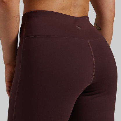 Close-up back view of a person wearing high-waisted leggings in a deep plum-brown color against a light gray studio background.
