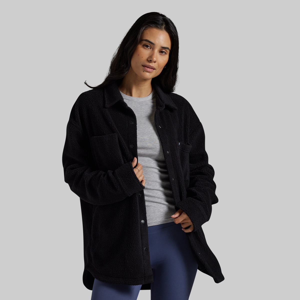 A woman modeling a black sherpa fleece button-up overshirt over a gray t-shirt and blue leggings, set against a solid light gray studio background.