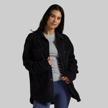 A woman modeling a black sherpa fleece button-up overshirt over a gray t-shirt and blue leggings, set against a solid light gray studio background.