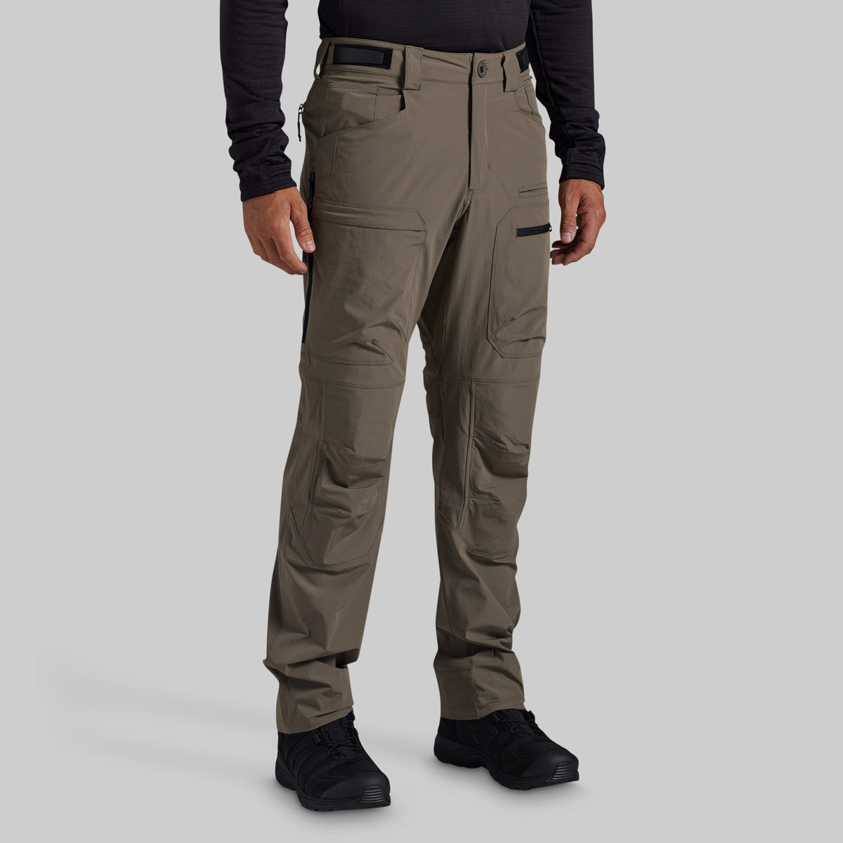 A person wearing grey outdoor cargo pants with black shoes and a black long-sleeve shirt, shown from the waist down against a light gray studio background.