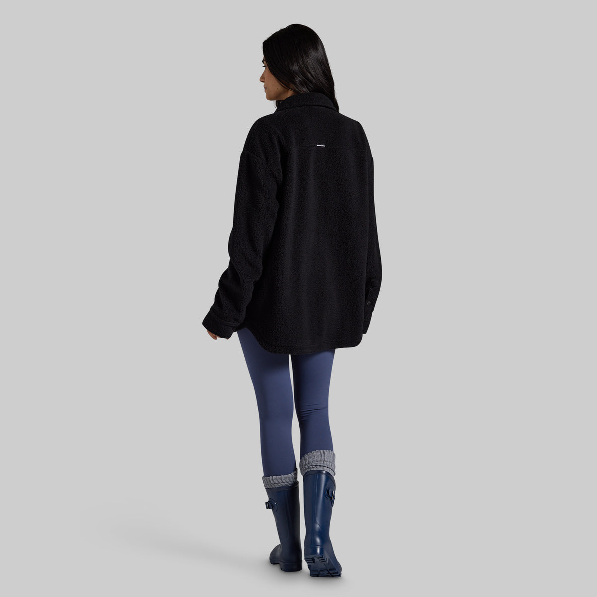 A woman is shown from the back wearing a black fleece jacket, blue leggings, and dark blue rain boots against a solid gray studio background.
