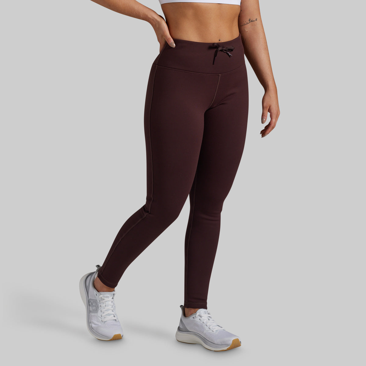 A model wearing high-waisted, ankle-length fleece lined leggings in a deep burgundy-brown color, paired with white sneakers, shown from the waist down against a solid light gray background.