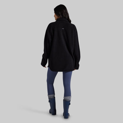 A woman is shown from behind wearing a black fleece pullover, blue leggings, and blue rain boots against a solid gray studio background.