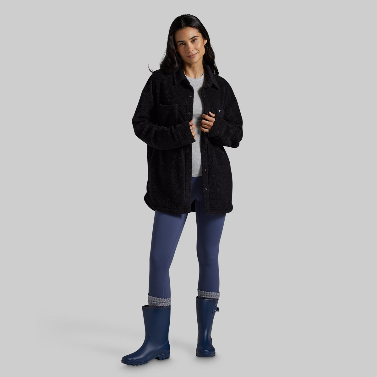 A woman modeling a black fleece button-up jacket over a white t-shirt, blue leggings, and blue rain boots, set against a solid gray studio background.