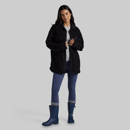 A woman modeling a black fleece button-up jacket over a white t-shirt, blue leggings, and blue rain boots, set against a solid gray studio background.
