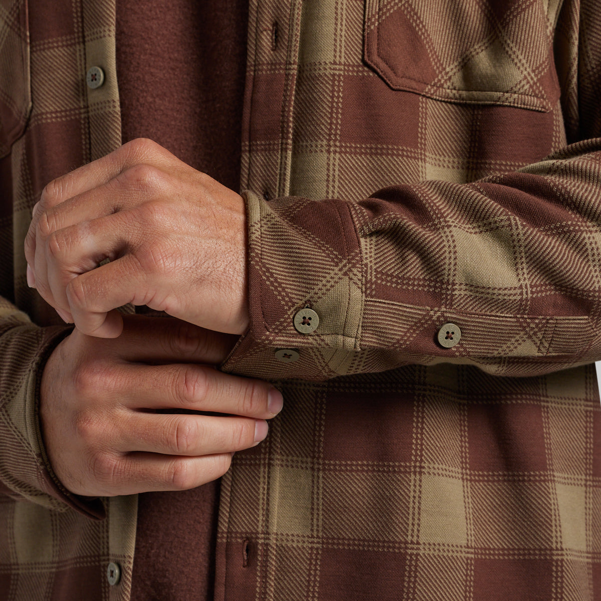 Close-up of a person wearing a brown and tan plaid flannel shirt, with both hands adjusting the cuff.