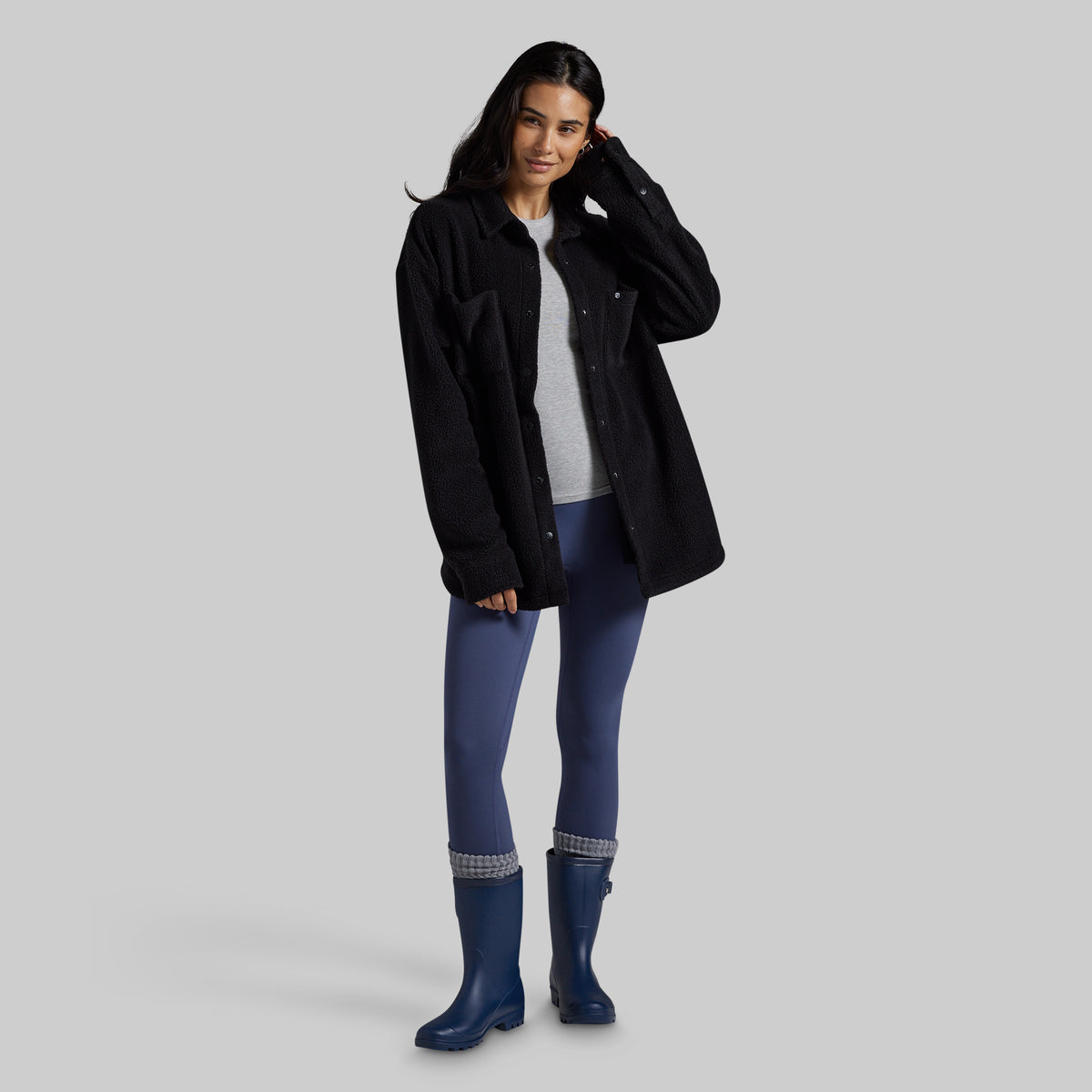 A woman modeling a black overshirt jacket over a light gray top, blue leggings, and blue rain boots against a solid gray studio background.
